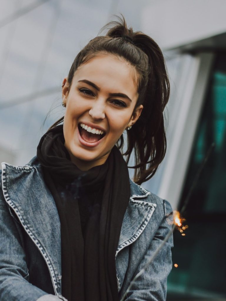 Lachende junge Frau mit Pferdeschwanz in Jeansjacke und Schal, vor unscharfem Hintergrund mit moderner Architektur.