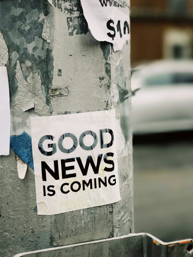 Plakat mit der Aufschrift 'GOOD NEWS IS COMING' an einem alten Laternenpfahl, umgeben von abgerissenen Zetteln, unscharfer Hintergrund.